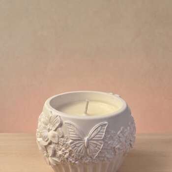 Vela artesanal Náyades en recipiente blanco con relieves de mariposas, flores y motivos naturales.