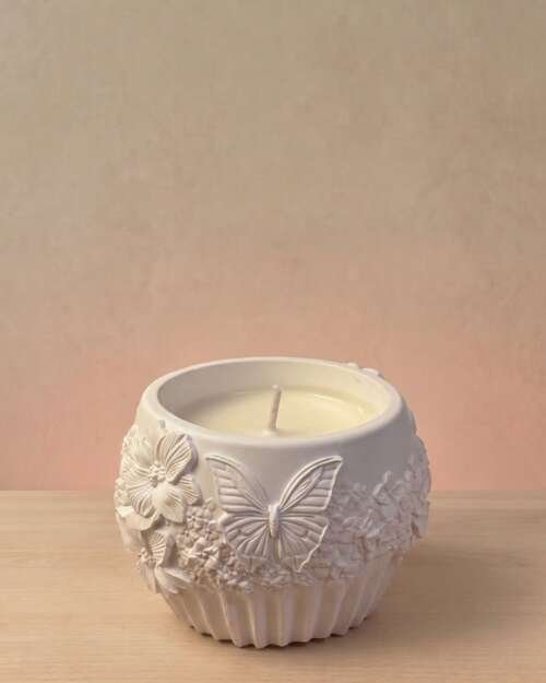 Vela artesanal Náyades en recipiente blanco con relieves de mariposas, flores y motivos naturales.