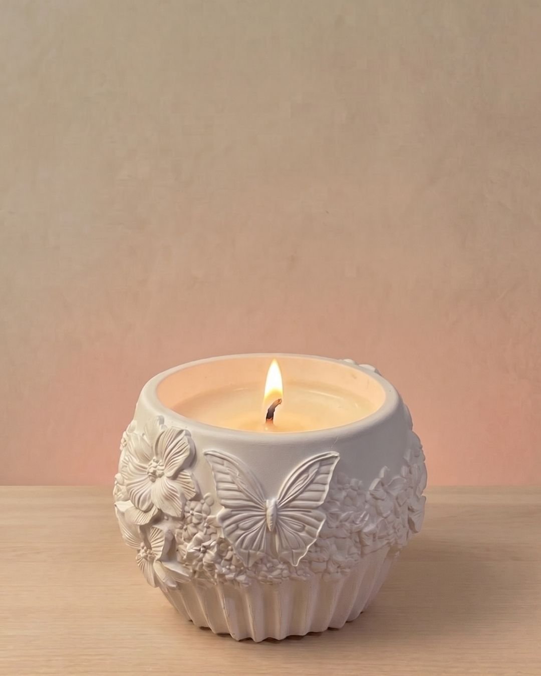 Vela artesanal Náyades en recipiente blanco con relieves de mariposas, flores y motivos naturales.