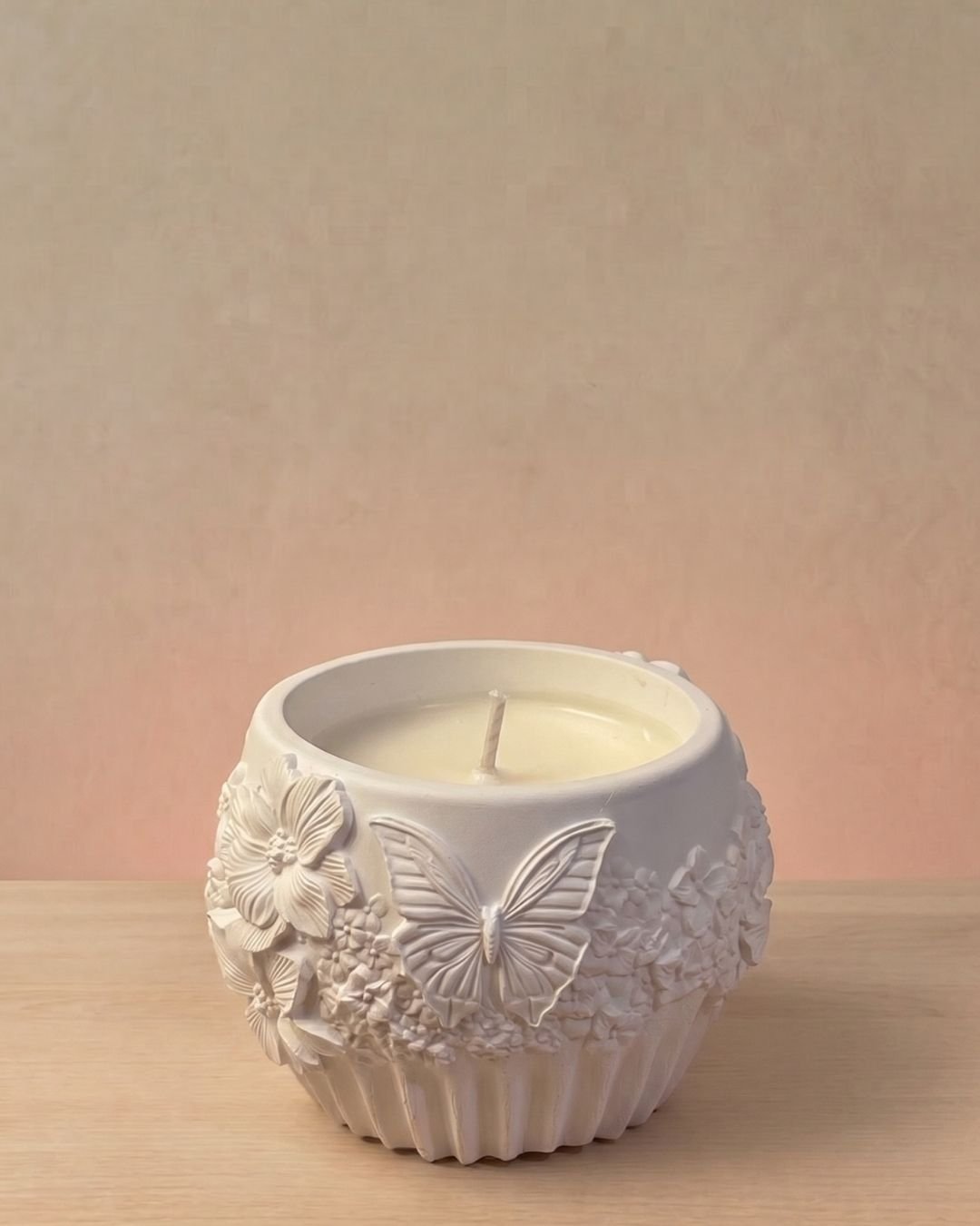Vela artesanal Náyades en recipiente blanco con relieves de mariposas, flores y motivos naturales.